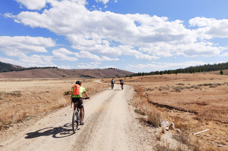 3 Riders from dirt drift pedaling away on a gravel road in an open plain.