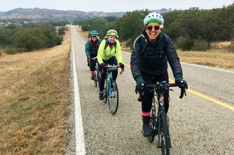 3 Riders smiling heading toward the camera on a hilly road with shrubs.