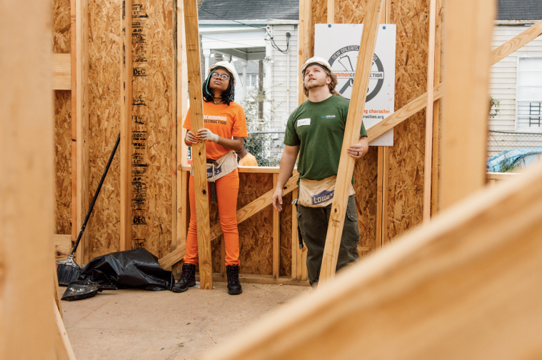 2 Volunteers with Uncommon Construction hold 2-inch by 6-inch lumber beams in the center of a build site.