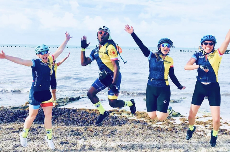 4 Riders from Keys to Canada off bicycles jumping on the beach.